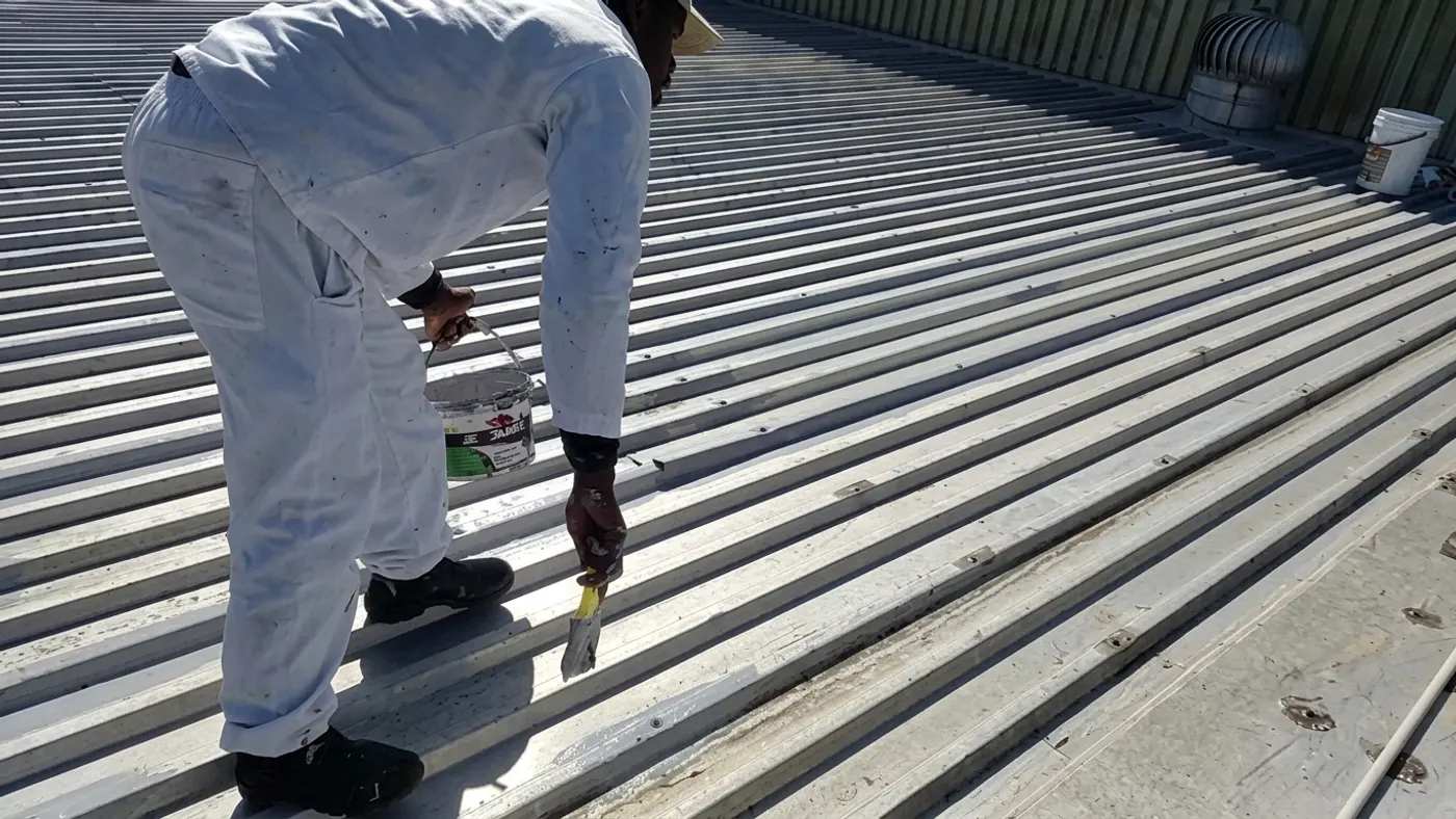 Roofing technician brush-applying white waterproof coating to a corrugated metal roof in Bellville